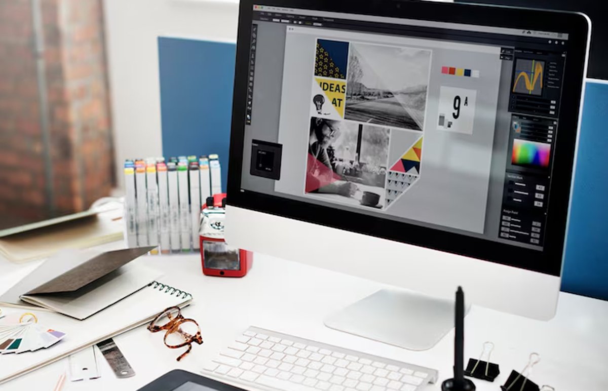 Services A high-resolution graphic designer's workspace featuring a large digital monitor displaying colorful brand identity layouts, social media ad creatives, and typography assets developed by Yuvox Marketing. On the desk sits a drawing tablet with a stylus, printed color swatches, and a sketchpad filled with initial logo concepts and UI design notes. The bright, collaborative office setting highlights professional visual storytelling, clean branding, and digital design in action.